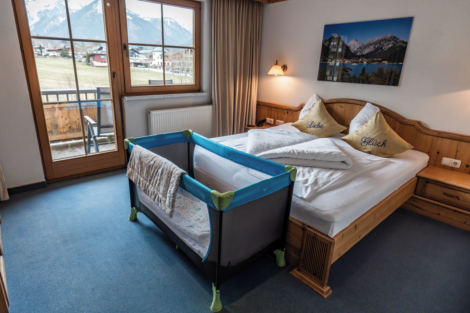 A room with a bed, a crib, a window with a view of mountains, a painting, and a lamp.