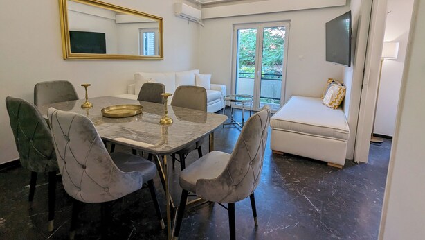 Uma sala de jantar com mesa de mármore, cadeiras estofadas, espelho com moldura dourada e um sofá com almofada estampada.