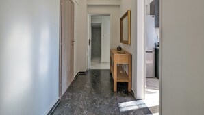 Um corredor com porta de madeira, uma cômoda de madeira com espelho e uma geladeira ao fundo.
