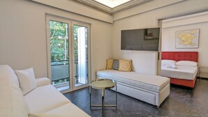 Quarto de hotel moderno com sofá branco, mesa redonda pequena, cama com cabeceira vermelha e uma grande janela com vista para área verde.