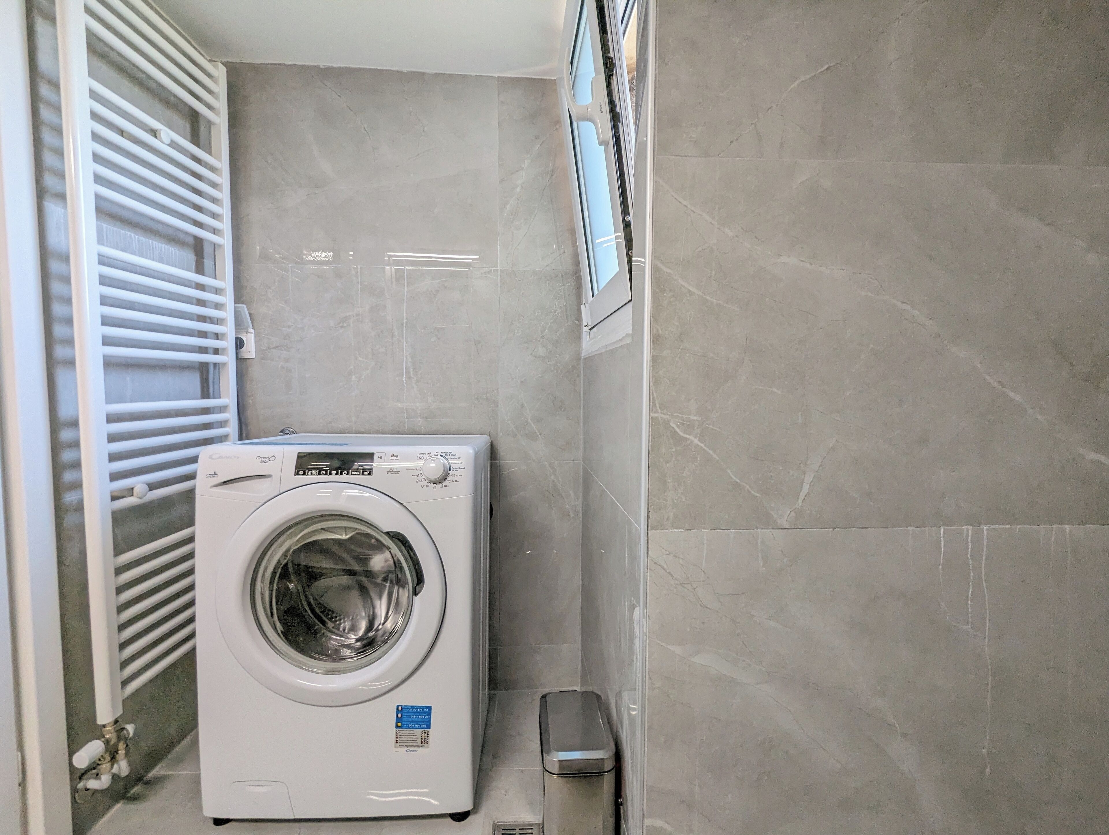 Un lavadero pequeño y moderno con lavadora de carga frontal, un cubo de basura y una ventana con persianas.