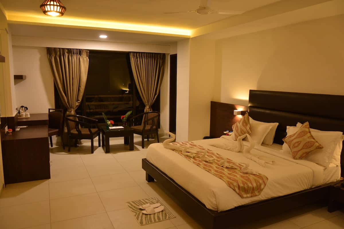 A hotel room with a large bed, a desk, and a chair. There is a window with curtains, a ceiling fan, and a nightstand with a lamp.