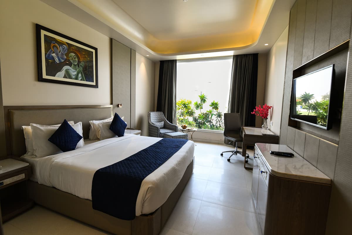 A hotel room with a large bed, a desk, a chair, a TV, and a window with a view of greenery.
