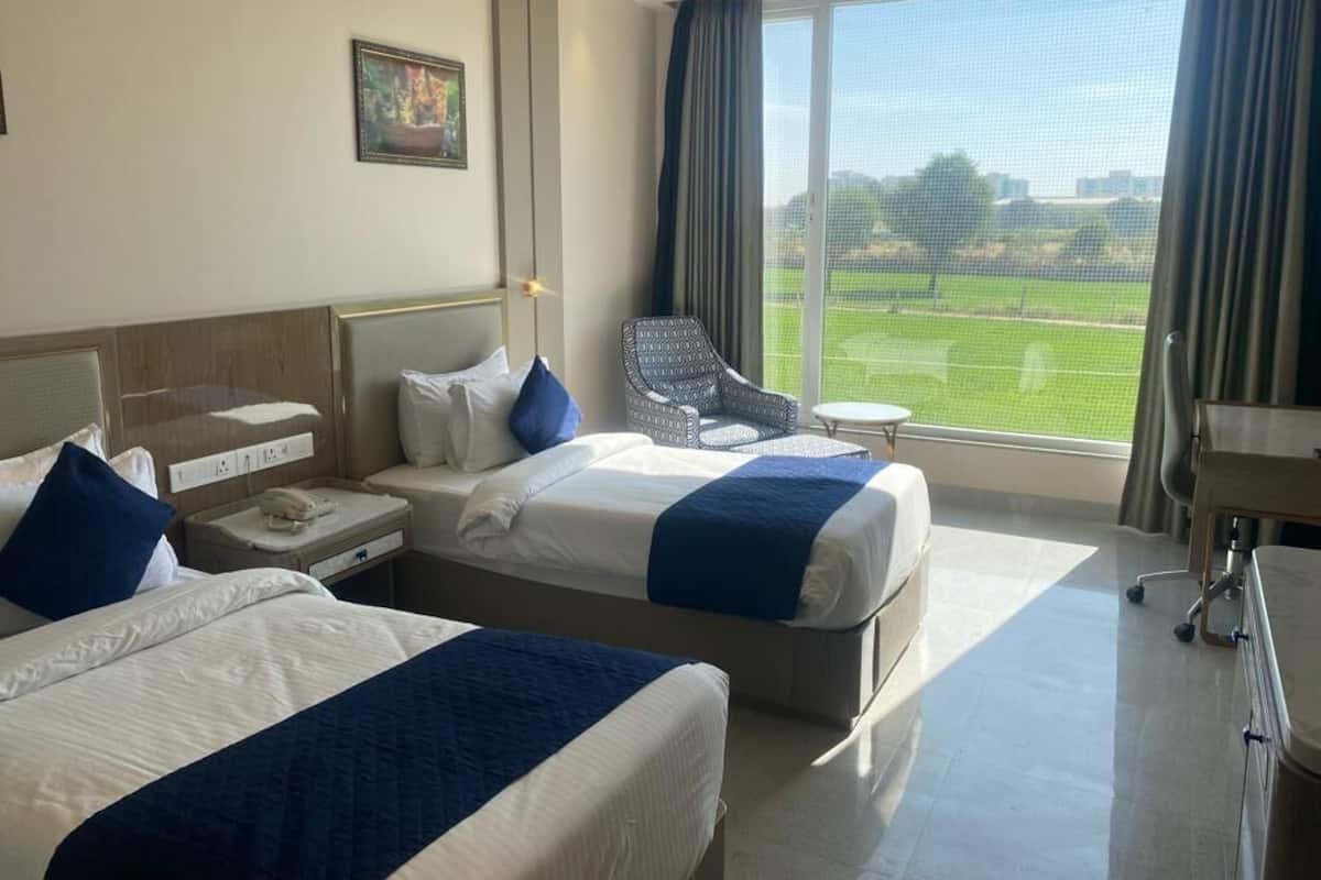 A hotel room with two beds, a chair, a small table, and a large window offering a view of a green field.