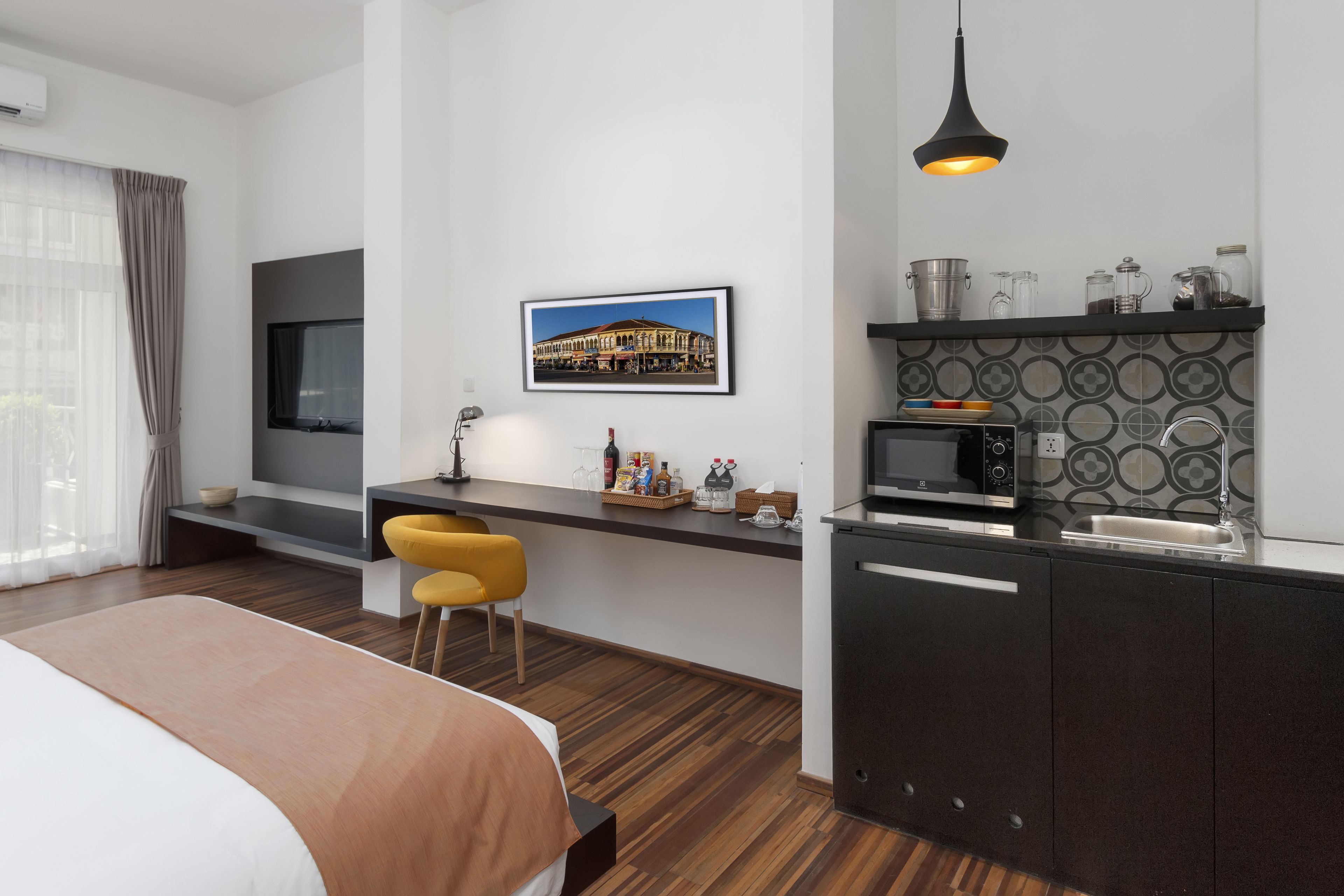 Una camera d'albergo moderna con un letto, una scrivania, una sedia, una TV e un angolo cottura con un forno a microonde, oltre a un quadro raffigurante un edificio.