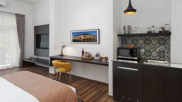Una camera d'albergo moderna con un letto, una scrivania, una sedia, una TV e un angolo cottura con un forno a microonde, oltre a un quadro raffigurante un edificio.