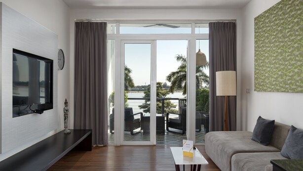 A modern living room with a flat-screen TV mounted on the wall, a sofa, a coffee table, and a view of the outdoors through large sliding glass doors. - Tao Riverside Residence (Phnom Penh)