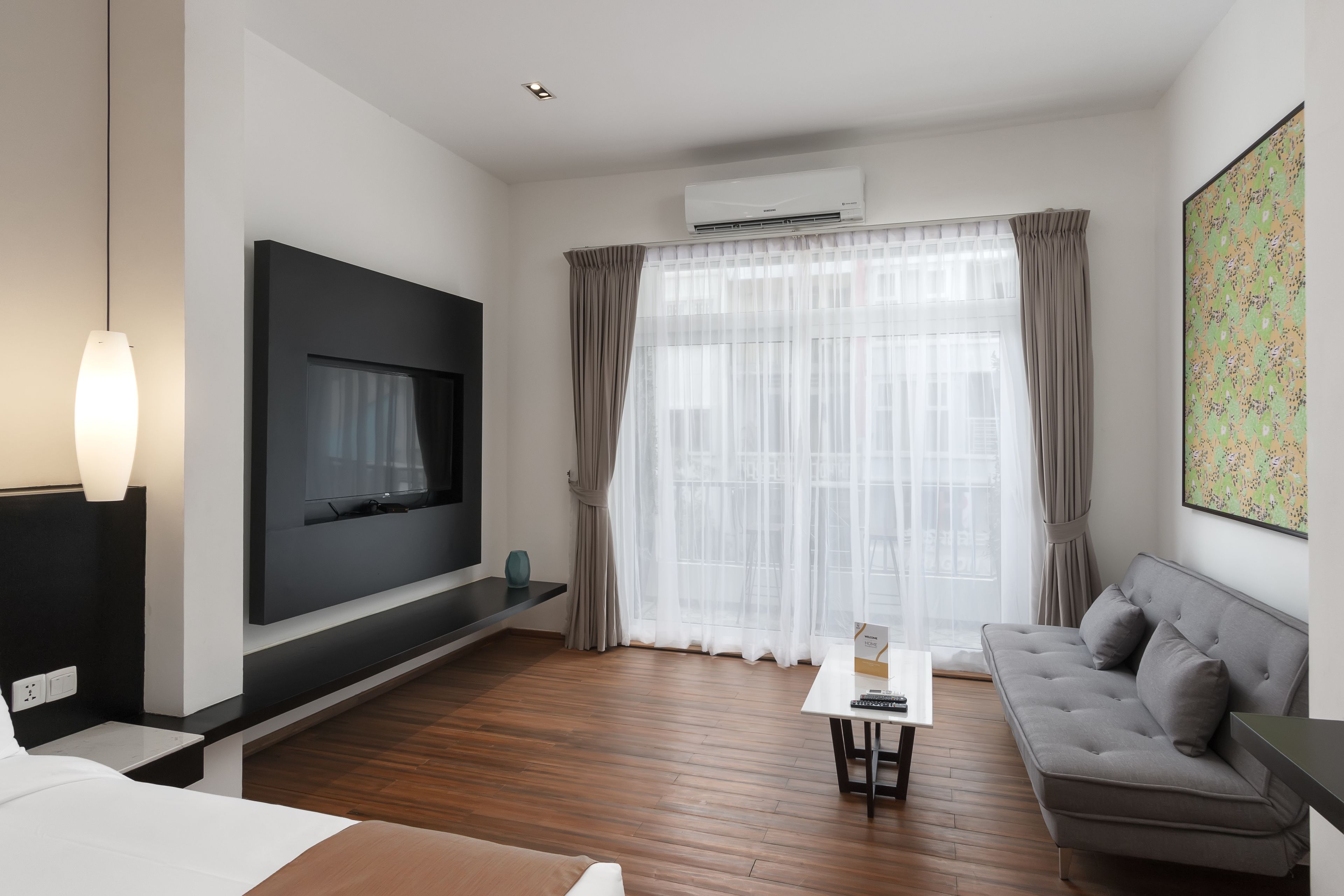 A modern hotel room with a flat-screen TV, a sofa, a small table, and a large window with sheer curtains.