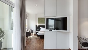 A modern hotel room with a large TV mounted on a white cabinet, a bed with white bedding, a small desk, and a chair. - Tao Riverside Residence (Phnom Penh)