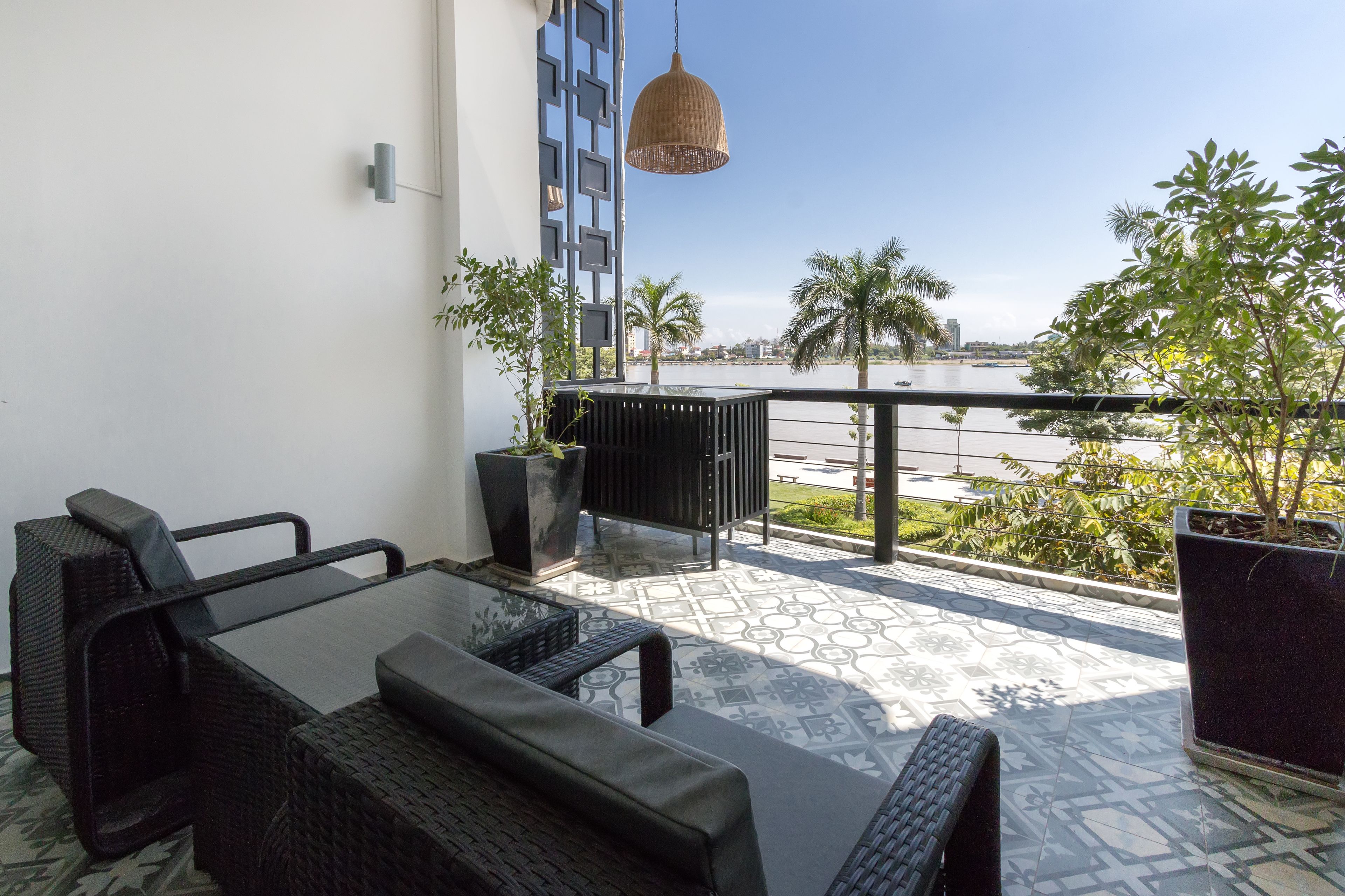 Ein Balkon mit Tisch und Stühlen, einer hängenden Leuchte, Topfpflanzen und Blick auf einen Fluss und Palmen.