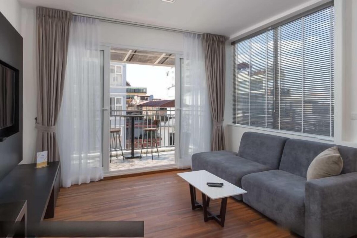 A modern living room with a grey sofa, a small white table, and a TV on a stand. There is a sliding glass door leading to an outdoor area with a view of buildings.