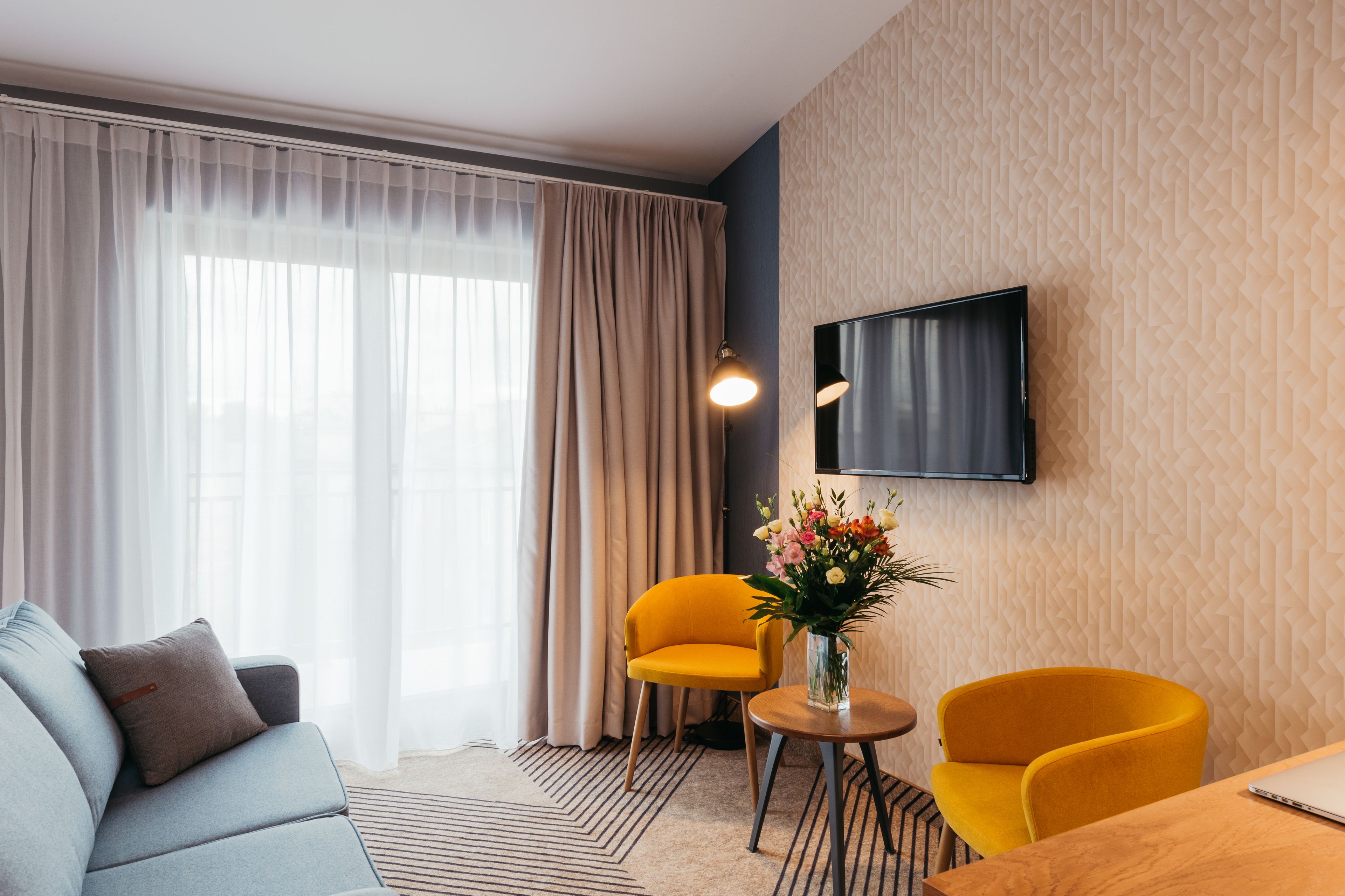 Una habitación de hotel moderna con sofá, dos sillas amarillas, una mesita con un jarrón de flores y un televisor de pantalla plana montado en la pared.
