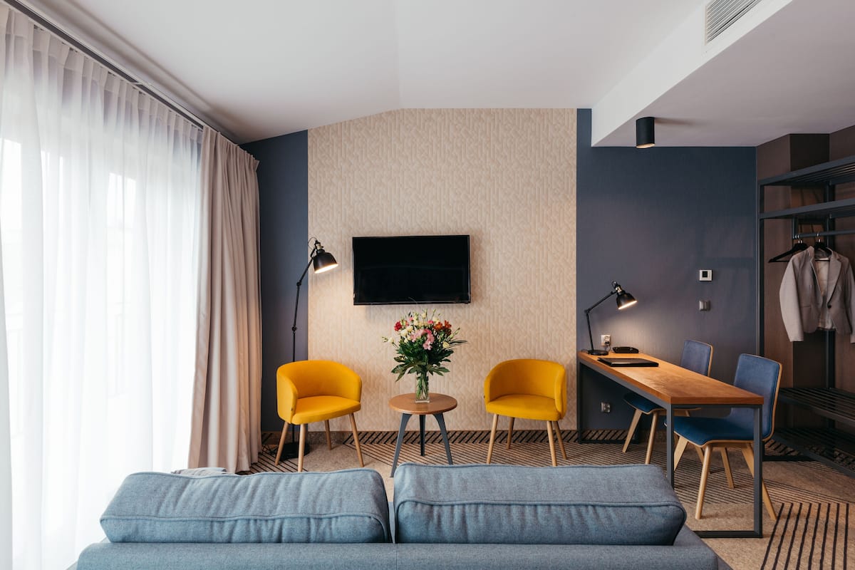 A modern hotel room with a sofa, two yellow chairs, a small table with a vase of flowers, a desk with a chair, a flat-screen TV, and a coat rack.