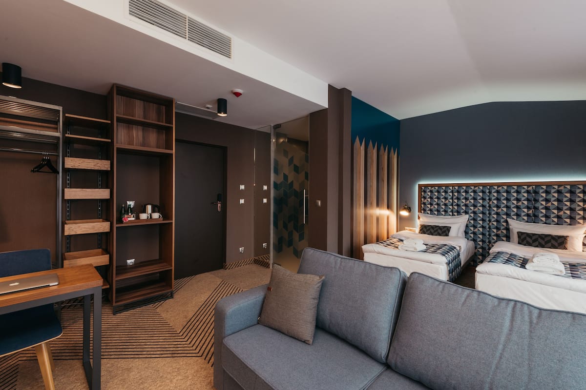 A modern hotel room with a grey sofa, two beds with patterned headboards, a wooden shelving unit, and a desk with a laptop.