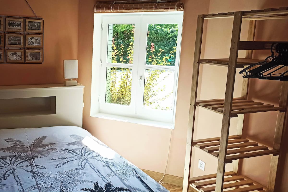 Quarto com cama, janela com vista para vegetação, prateleira na parede com quadros e um suporte de madeira.