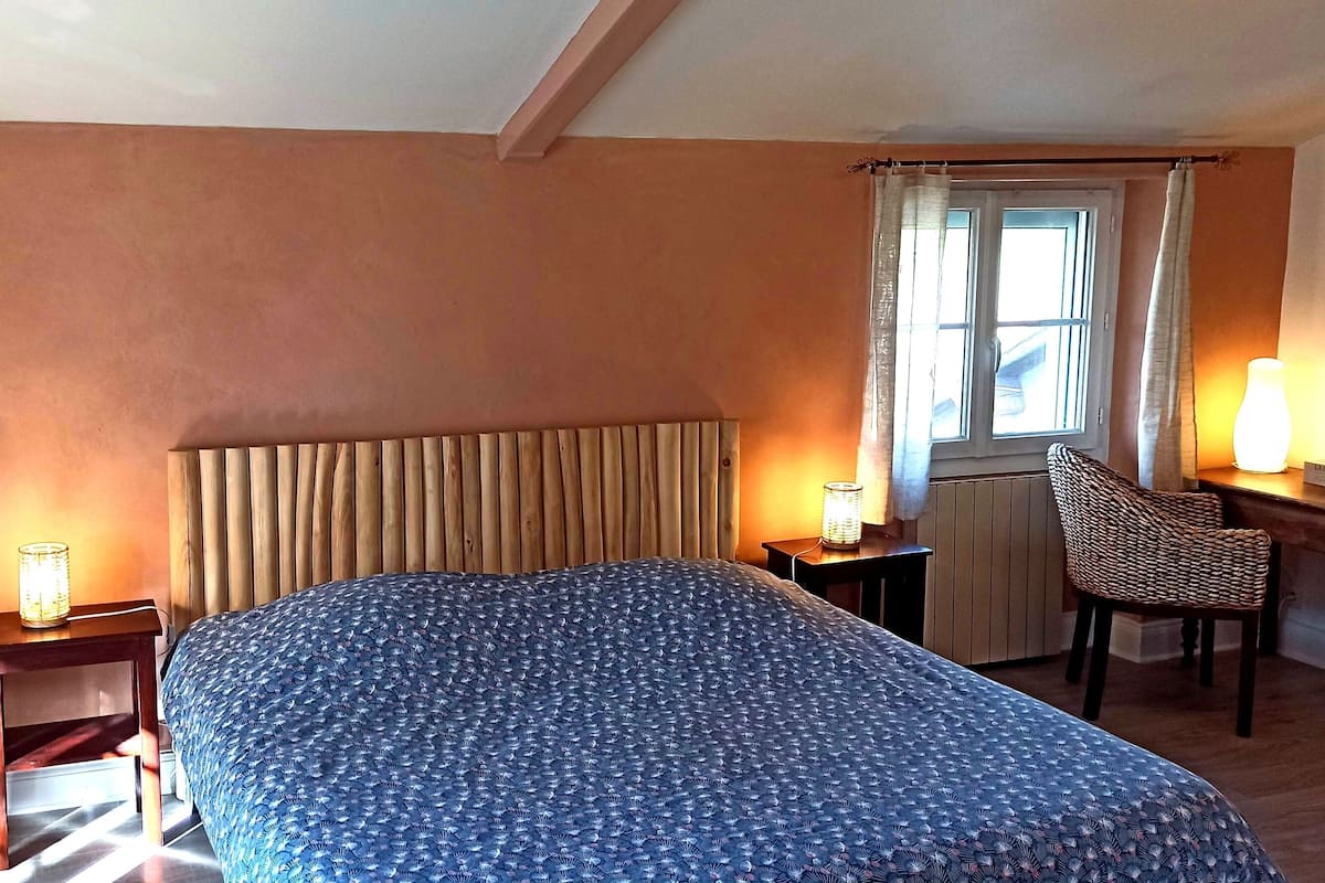 Quarto com cama, cadeira, mesa de cabeceira, luminária e janela com cortinas.