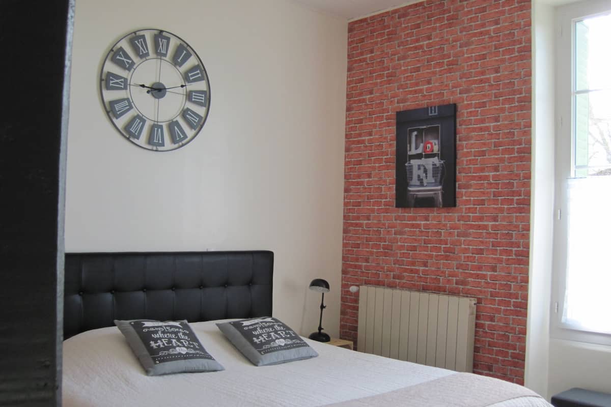 Quarto com cabeceira preta, duas almofadas com texto, relógio de parede, parede de tijolos e uma janela.