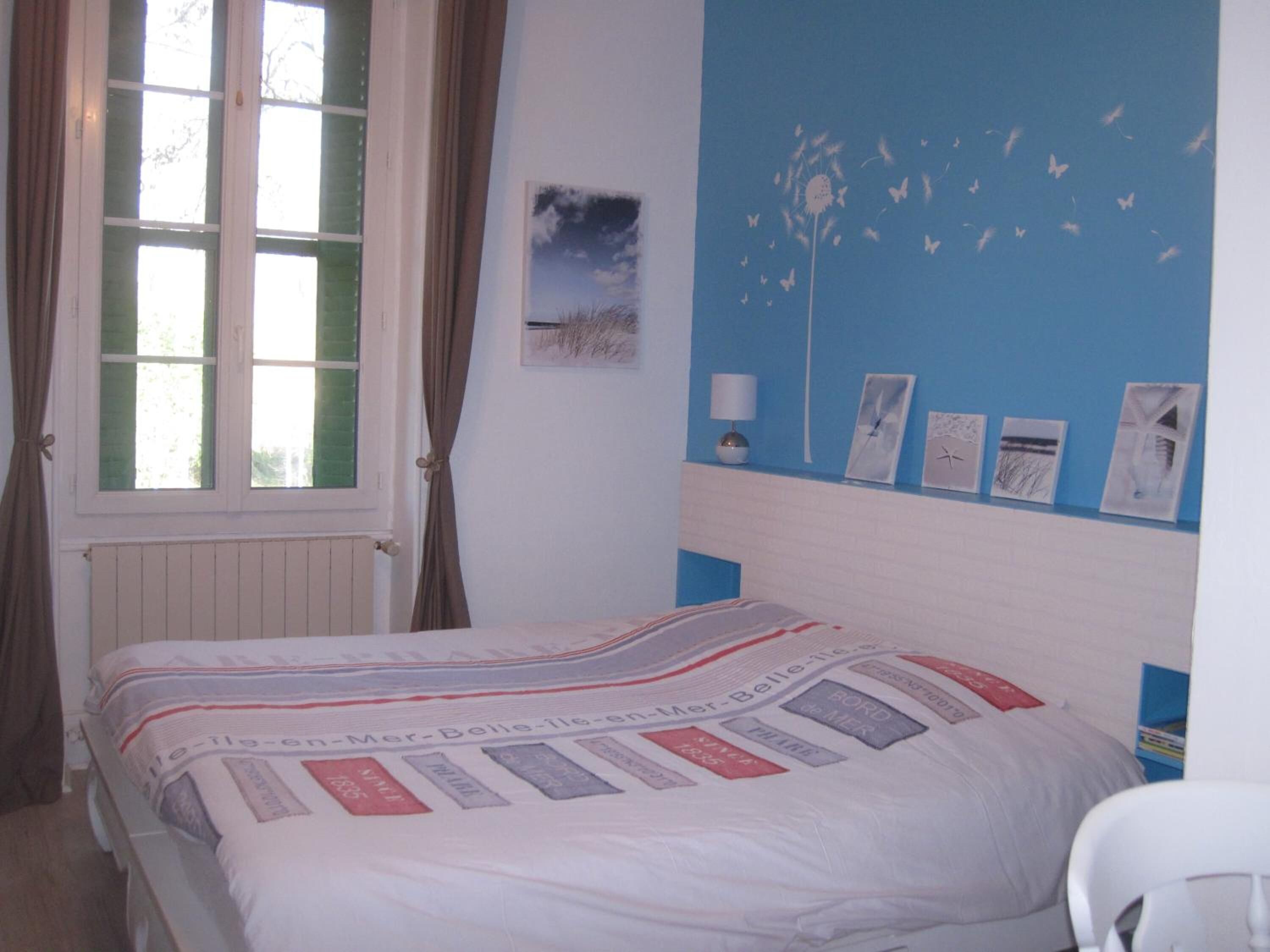 A bedroom with a bed, a window with curtains, a wall with framed pictures, and a blue accent wall.