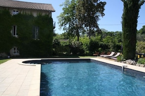 A swimming pool with a tiled deck, surrounded by lush greenery and a large tree. - Chambre d'hôtes Park des Collines - Gîte (Genissieux)