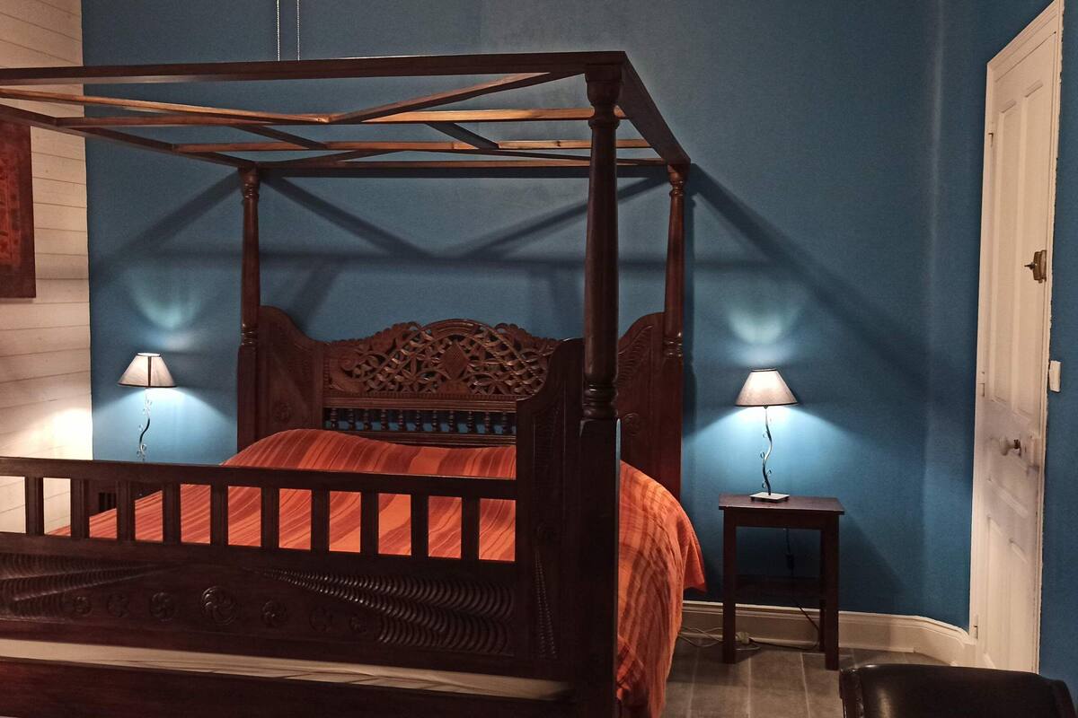 Uma cama de dossel com cabeceira de madeira, duas luminárias de cabeceira e uma mesinha ao lado.