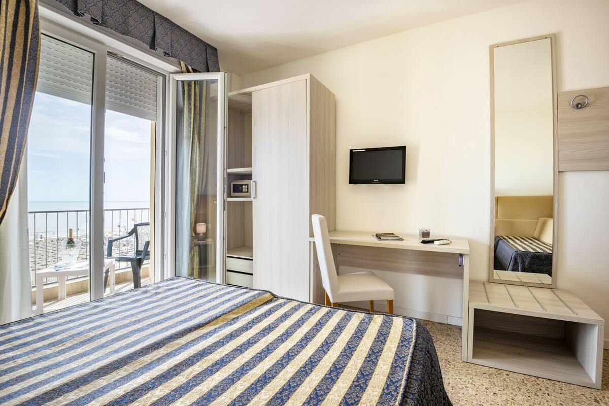 Habitación de hotel con cama, escritorio, silla, televisor y balcón con vista a la playa.