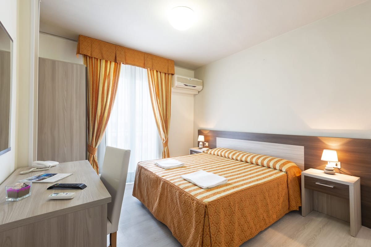Habitación de hotel con cama, mesitas de noche, un escritorio con silla, lámpara y ventana con cortinas.