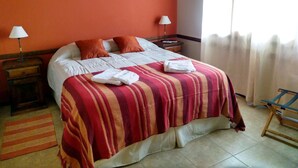 A neatly made bed with a striped bedspread, two pillows, and a folded towel on the bed. There are two bedside tables with lamps, a window with curtains, and a chair. - Alquimia Cabañas Apart (El Bolson)