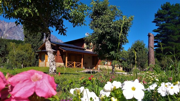 A rustic wooden cabin with a sloped roof, surrounded by lush greenery and flowering plants. - Alquimia Cabañas Apart (El Bolson)