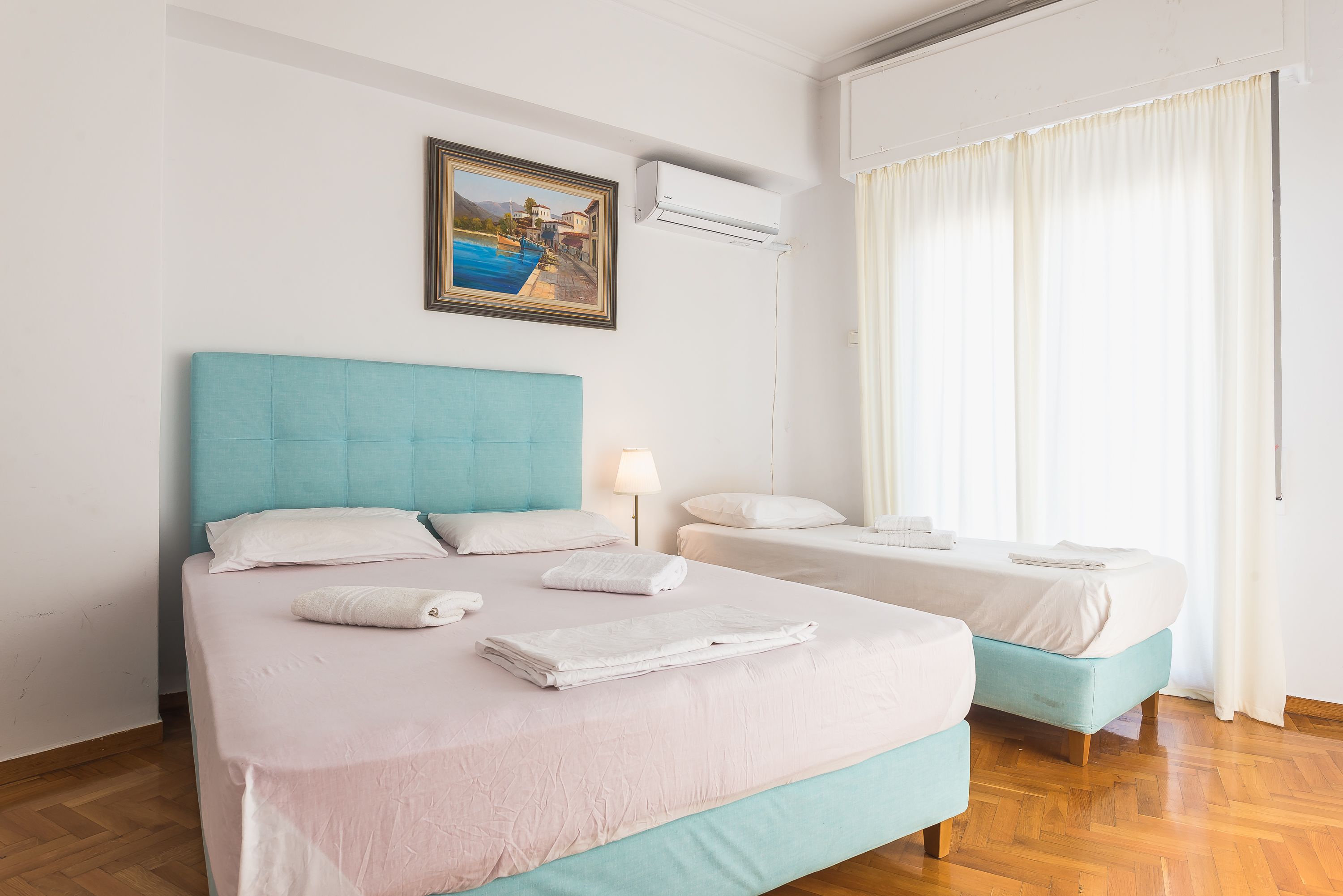 A double bed with a teal headboard, a bedside table with a lamp, and a painting of a coastal scene.