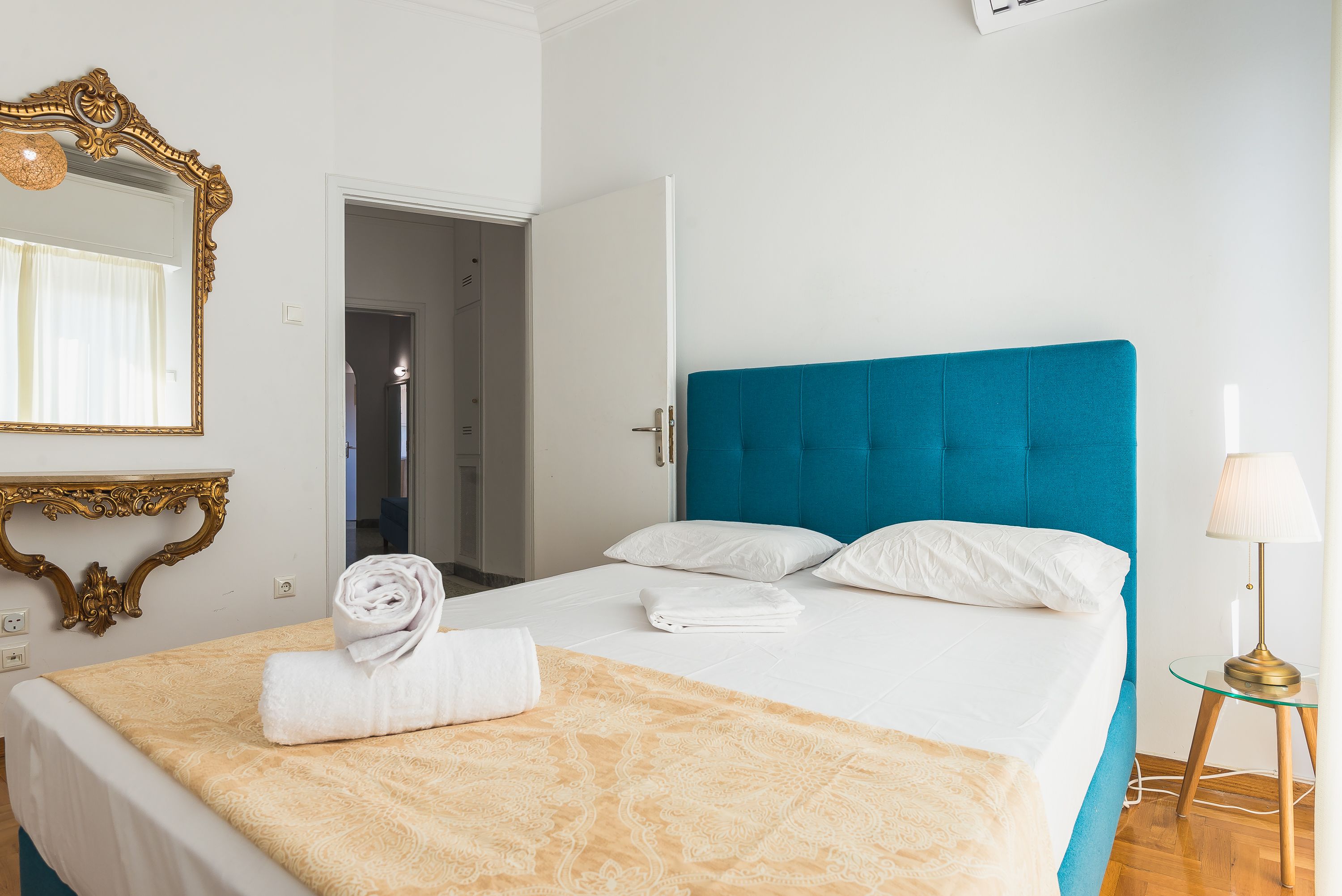 A hotel room with a bed, a mirror, a bedside table with a lamp, and a door leading to another room.