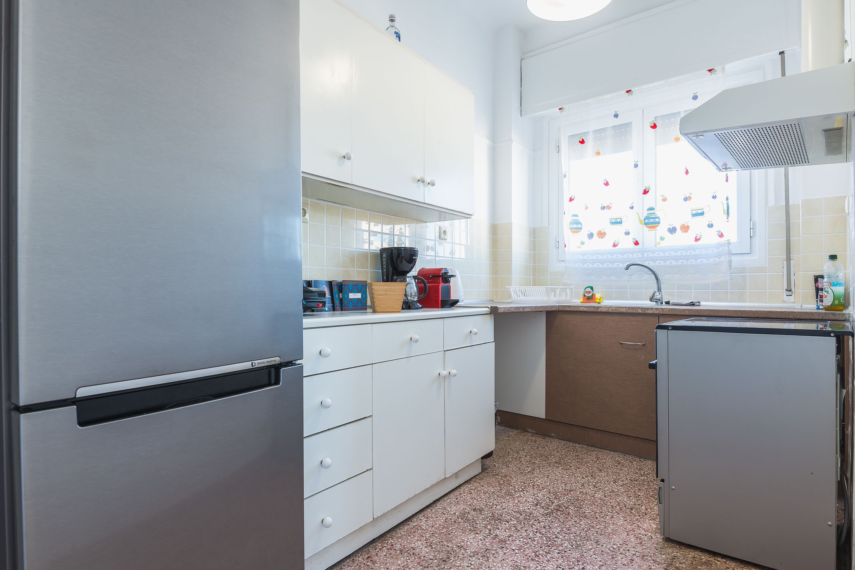 Eine kompakte Küche mit weißen Schränken, einem Edelstahlkühlschrank, einer Kaffeemaschine und einem Fenster mit Obstmuster.