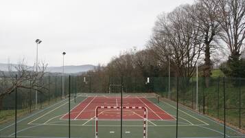 An outdoor basketball court with a red and green playing surface, surrounded by a chain-link fence and trees.