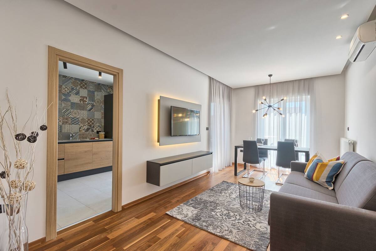 Ein modernes Wohnzimmer mit einem an der Wand befestigten Flachbildfernseher, einem grauen Sofa mit bunten Kissen, einem Holzboden und einem Essbereich im Hintergrund.