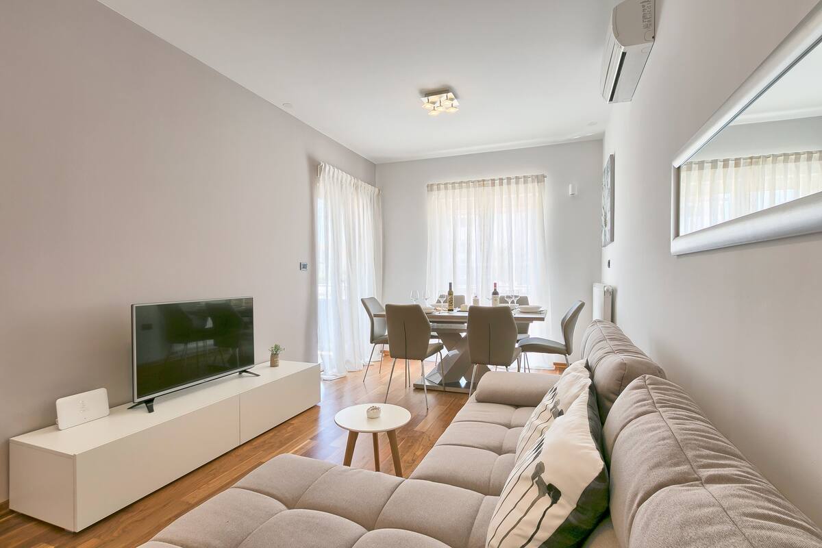 Ein modernes Wohnzimmer mit einem Flachbildfernseher auf einer weißen Konsole, einem beigefarbenen Sofa und einem Essbereich mit Tisch und Stühlen.