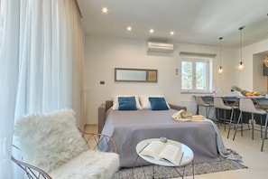 Ein modernes Schlafzimmer mit einem großen Bett, einem kuscheligen Sessel, einem kleinen Tisch mit einem Buch und einem Essbereich mit Stühlen und einer Obstschale.