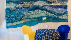 Um quarto com uma grande tapeçaria na parede, uma cadeira amarela, uma cadeira azul e uma mesa com toalha quadriculada.