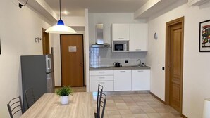 Uma cozinha compacta com armários brancos, micro-ondas e pia. Há uma mesa de jantar com cadeiras e uma pequena planta.