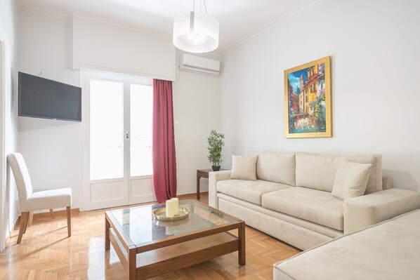 A living room with a sofa, a coffee table, a chair, a TV, and a painting on the wall. - Victoria Best Apartments (Athens)
