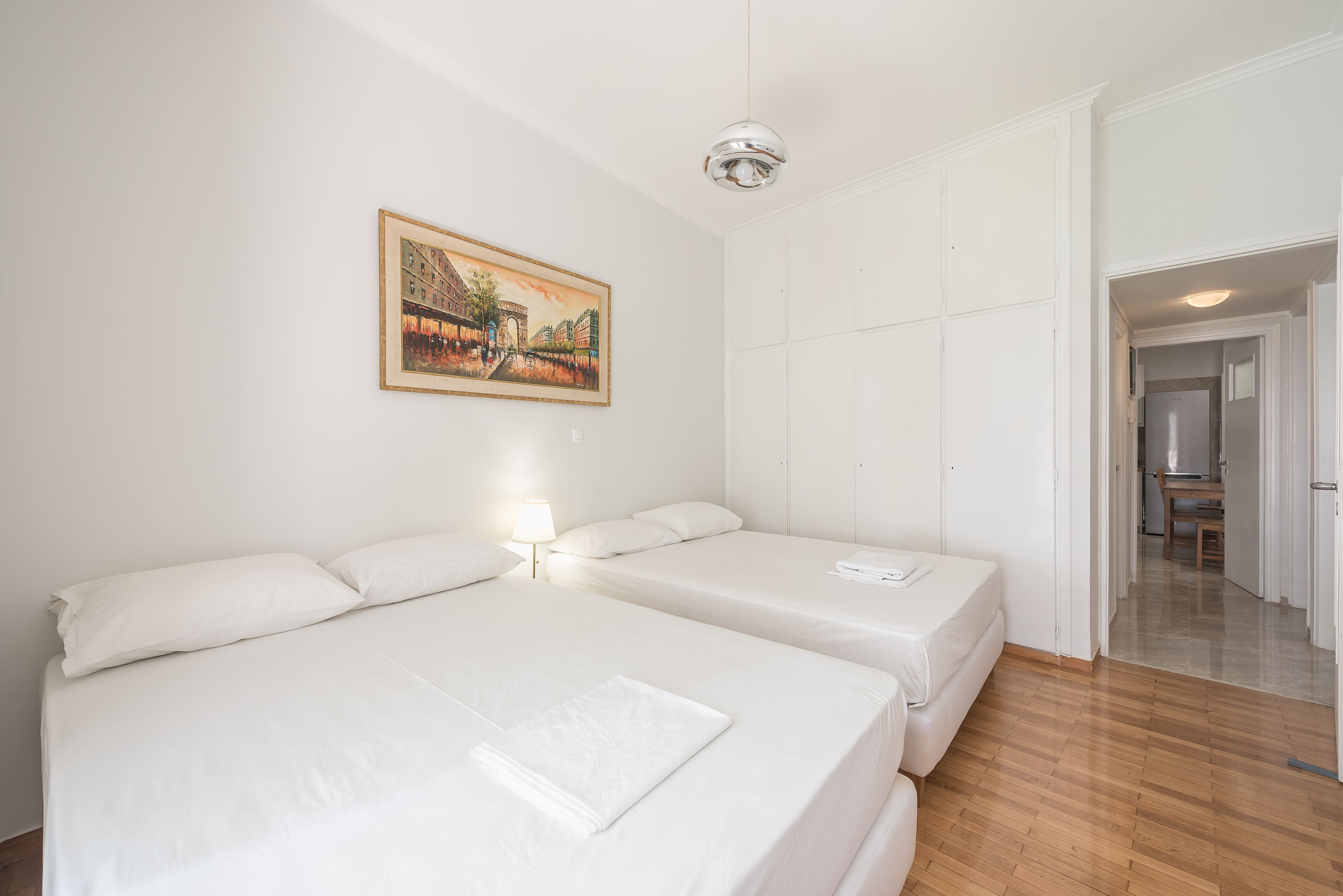 A hotel room with two beds, a painting on the wall, and a hallway leading to a dining area.
