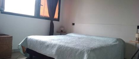 A single bed in a room with a window, a small table with a lamp, and a wicker basket.