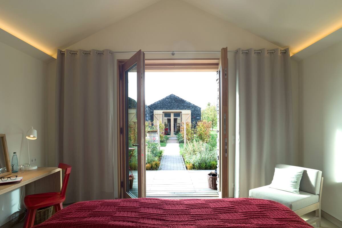 Un dormitorio con cama, escritorio, silla y vista a un jardín a través de una puerta abierta.