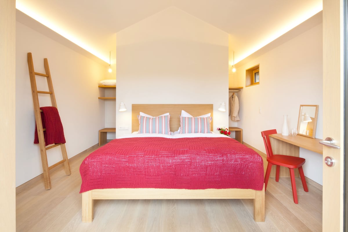 Un dormitorio con cama, una silla, un escritorio, una escalera y una ventana pequeña.