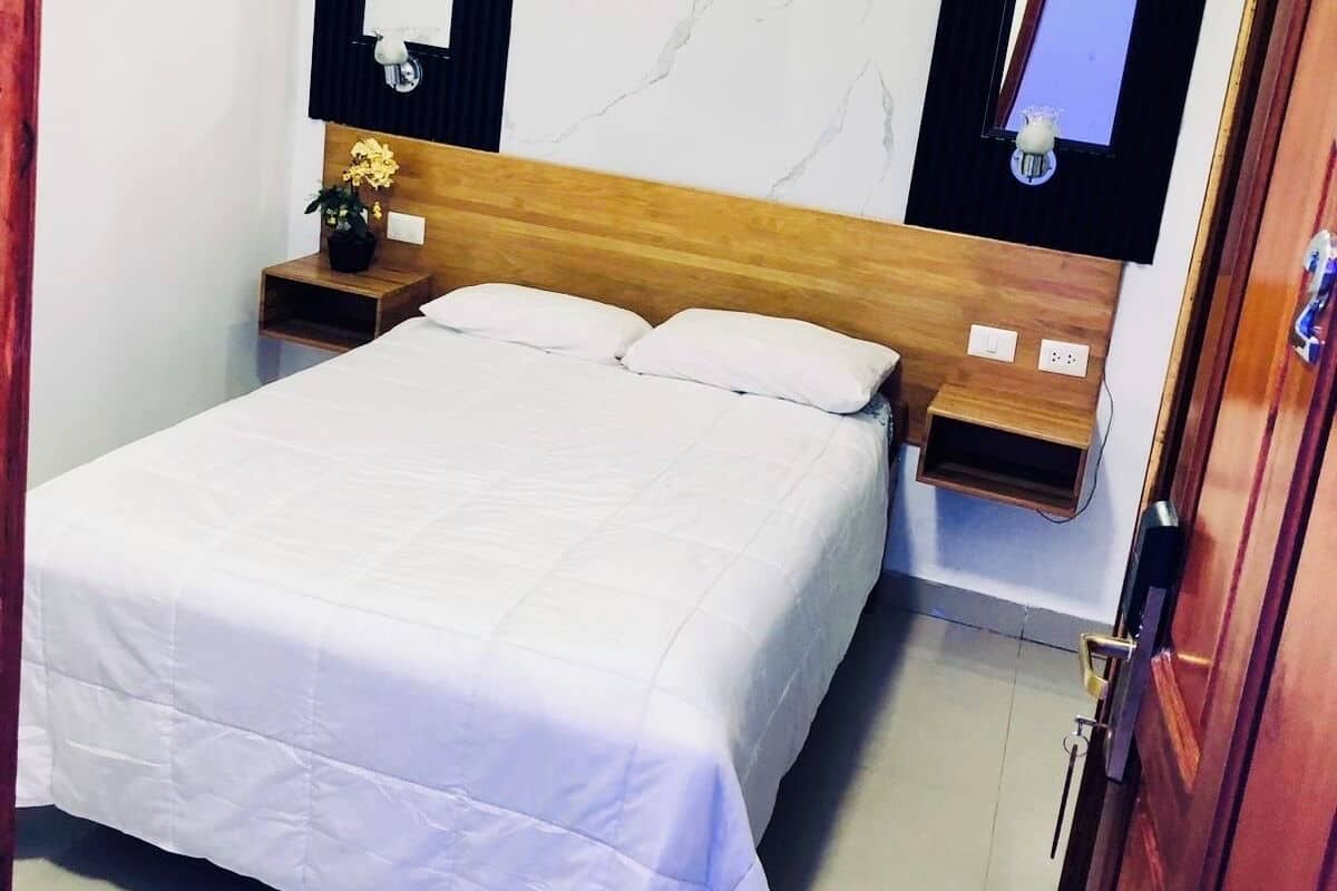 A neatly made bed in a hotel room with a marble wall, a mounted air conditioner, and a small bedside table with a lamp.
