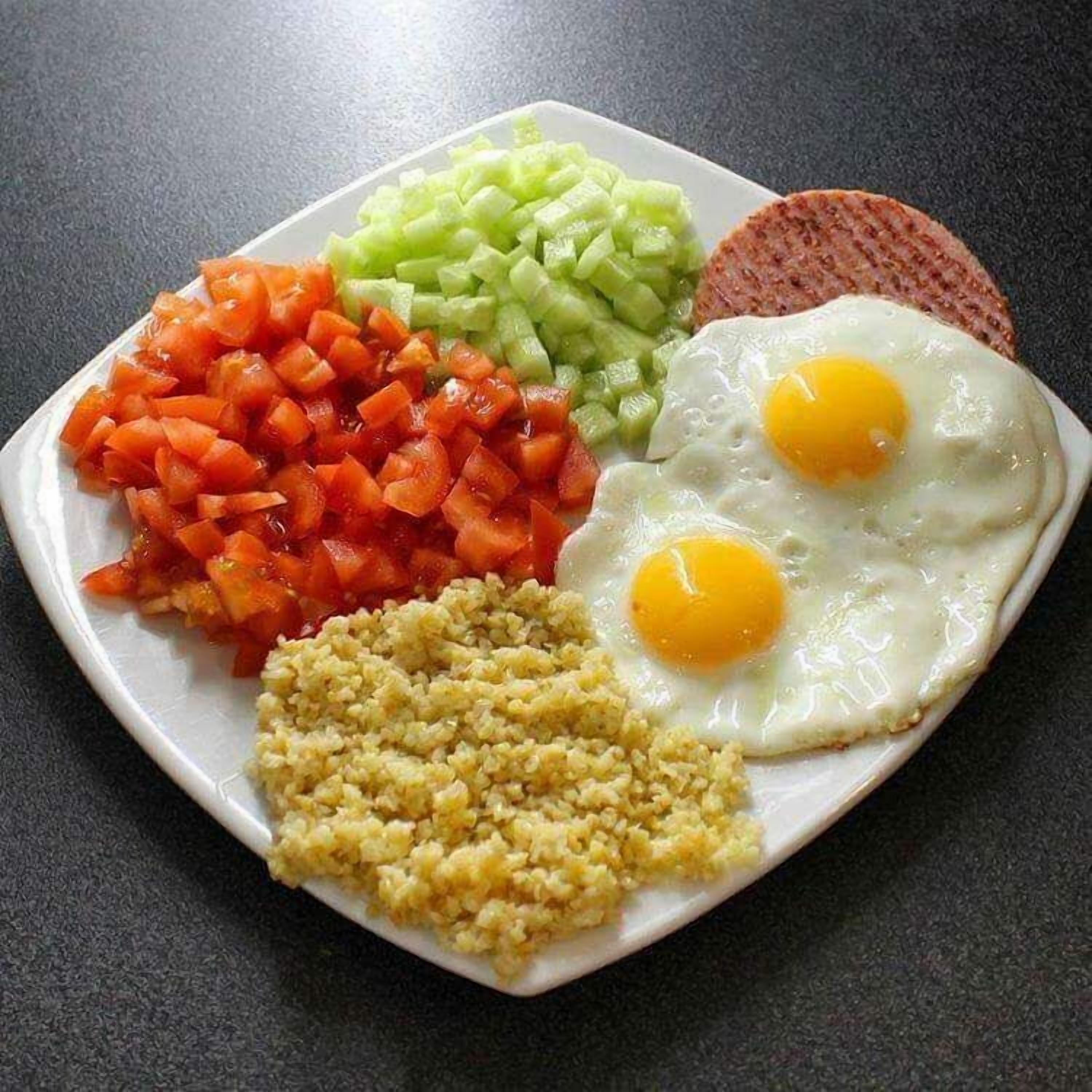 Ein Teller mit zwei Spiegeleiern, gewürfelten Tomaten, gewürfelten Gurken und einer Portion Couscous.