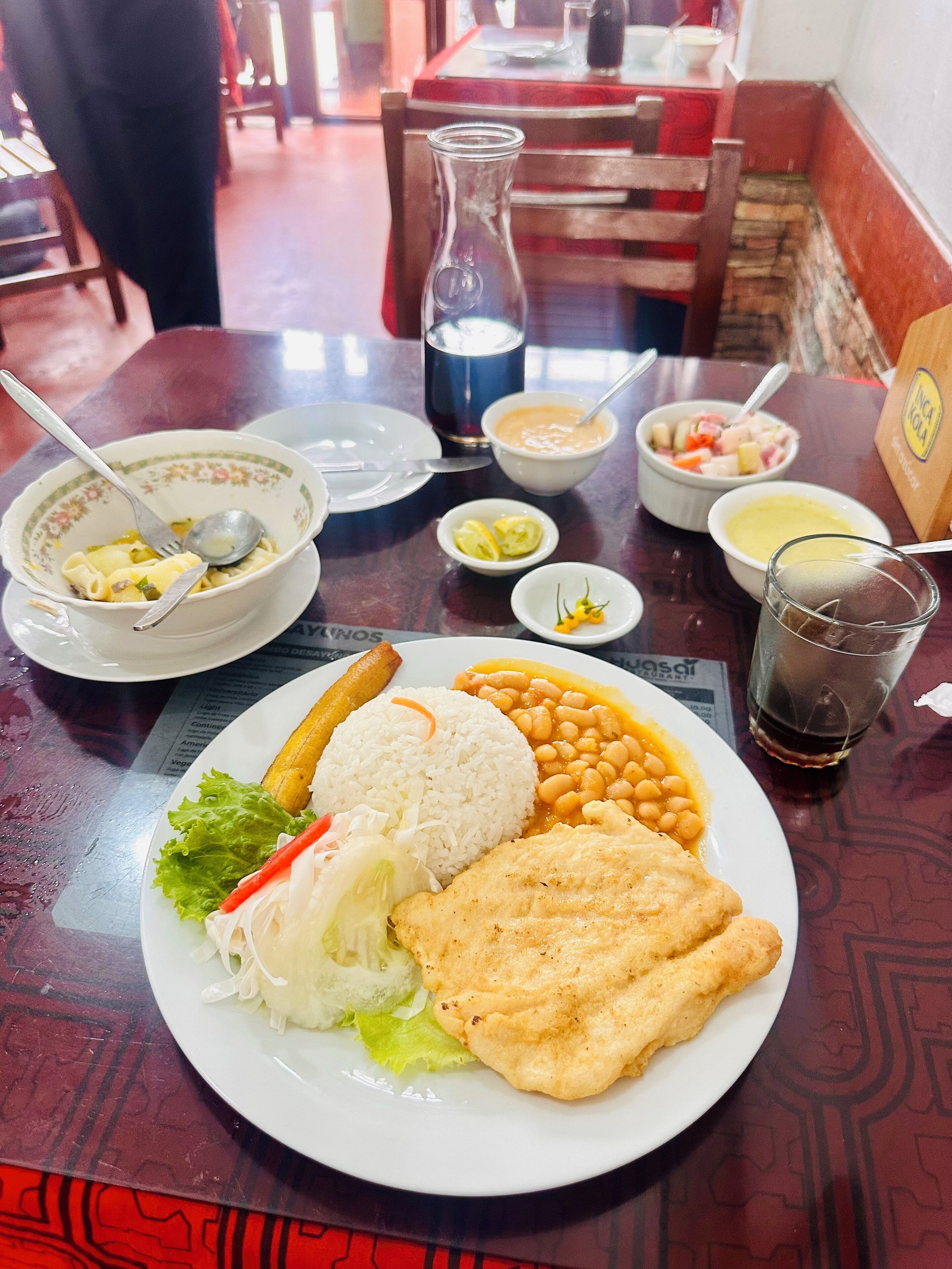 Una mesa de comedor con un plato de comida, que incluye arroz, frijoles y un alimento frito, junto con tazones de sopa y un vaso de refresco.
