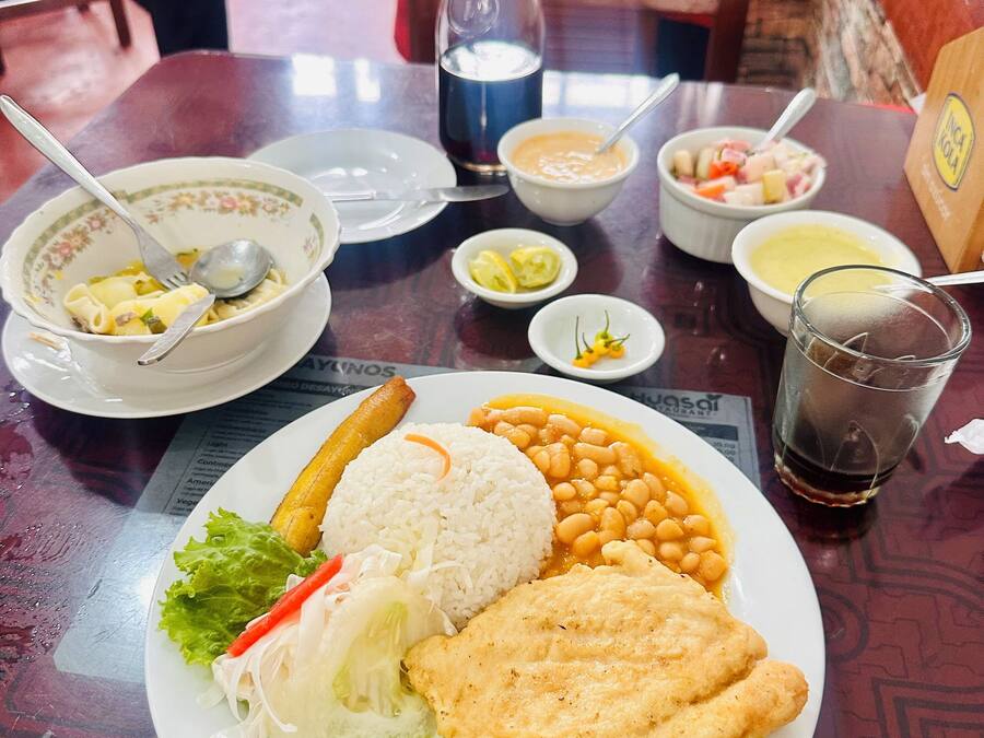 Une table à manger avec une assiette de nourriture, comprenant du riz, des haricots et un plat frit, ainsi que des bols de soupe et un verre de soda.