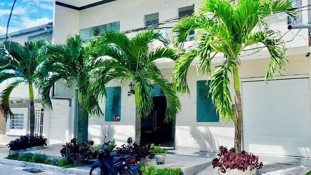 A modern building with a white facade, multiple windows, and a garage door. There are several palm trees and potted plants in the front yard. - Hotel Fenix (Iquitos)