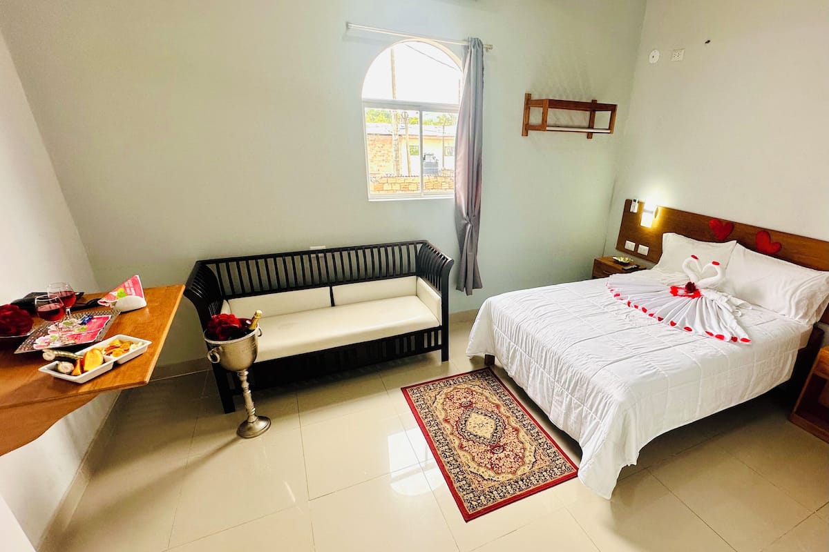 A hotel room with a bed, a sofa, a small table with food and drinks, and a window with curtains.