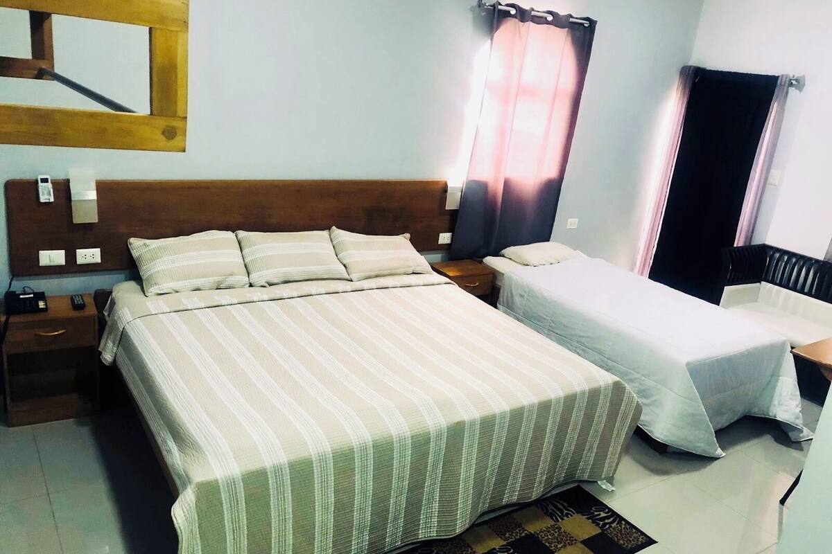 A hotel room with a large bed, a smaller bed, a nightstand, a desk, a chair, a window with curtains, and a mirror on the wall.