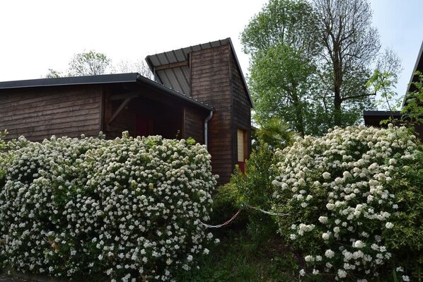 A rustic wooden cabin with a sloped roof, surrounded by dense clusters of white flowering bushes. - Relais des Cinq Routes (Parnac)